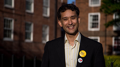 Dave Raval smiling and looking straight ahead. Blocks of redbrick flats in the background